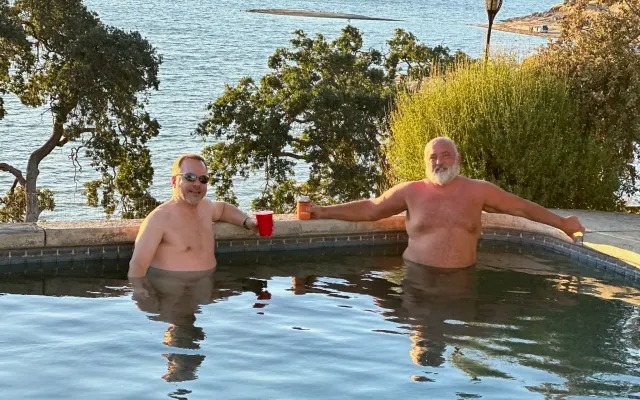 Two men toasting with drinks in the pool at golden hour, Lake Millerton behind them